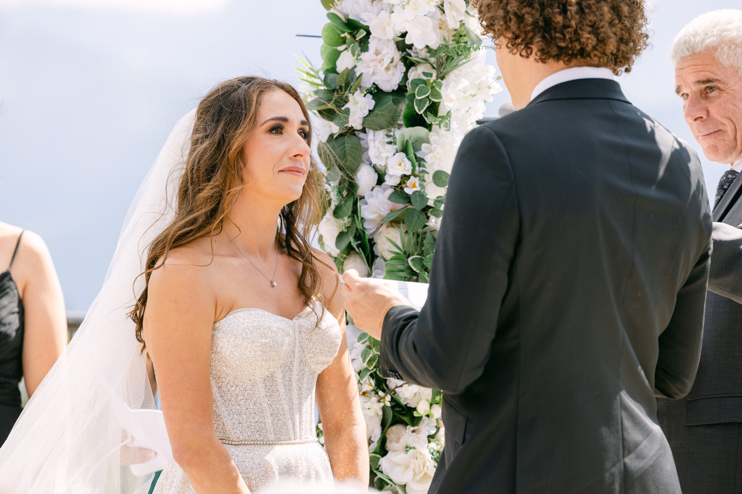 A bride listening intently during her wedding vows, with floral decorations in the background.