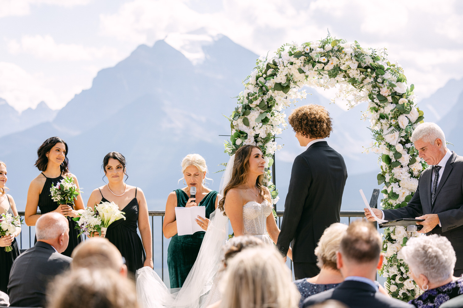 A couple exchanges vows in front of a floral arch amidst a stunning mountain backdrop, surrounded by wedding guests and bridesmaids.