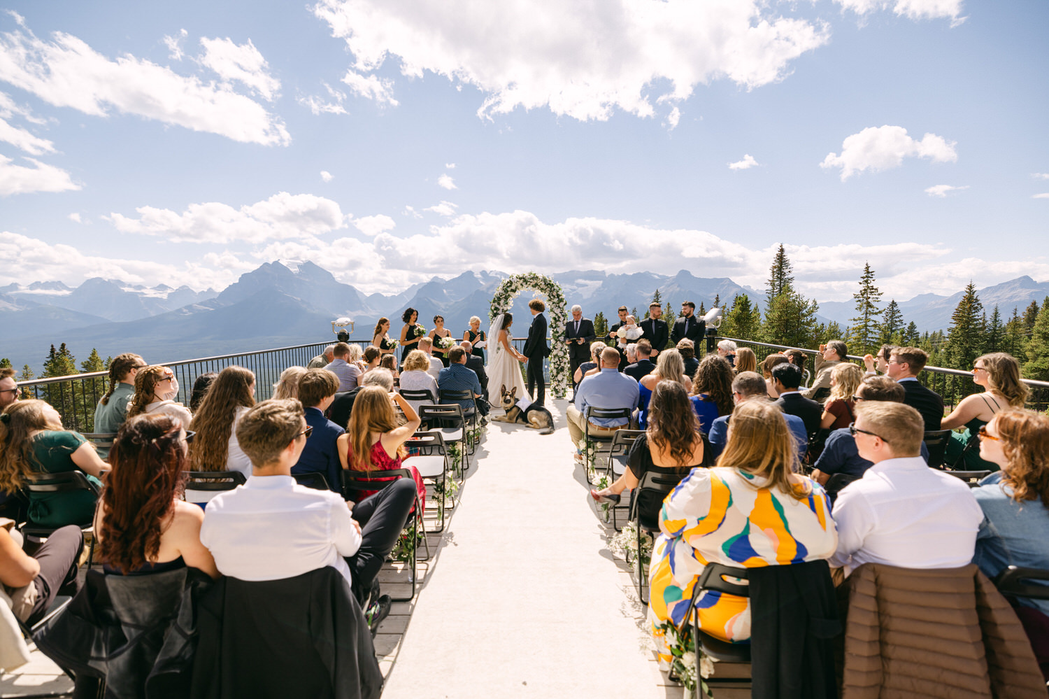 A scenic outdoor wedding ceremony with guests seated on a terrace, surrounded by mountains and trees, as the couple exchanges vows under a floral arch.