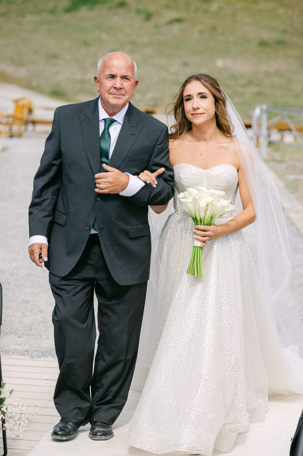 A bride in a shimmering gown walks down the aisle, accompanied by her father, both smiling amidst a scenic backdrop.