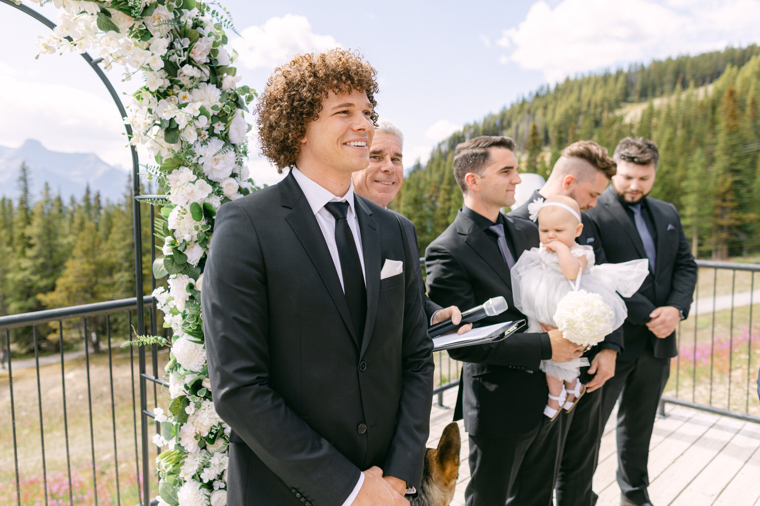 A groom smiles while standing before an arch adorned with flowers, surrounded by groomsmen and a baby, set against a mountainous landscape.