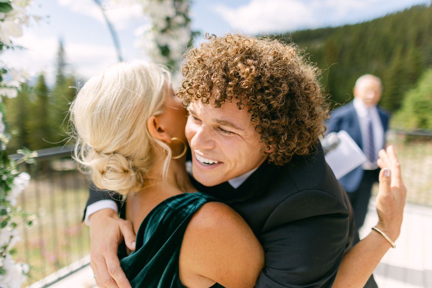 A man with curly hair shares a warm hug with a woman in a green dress, set against a picturesque outdoor backdrop with trees and flowers.