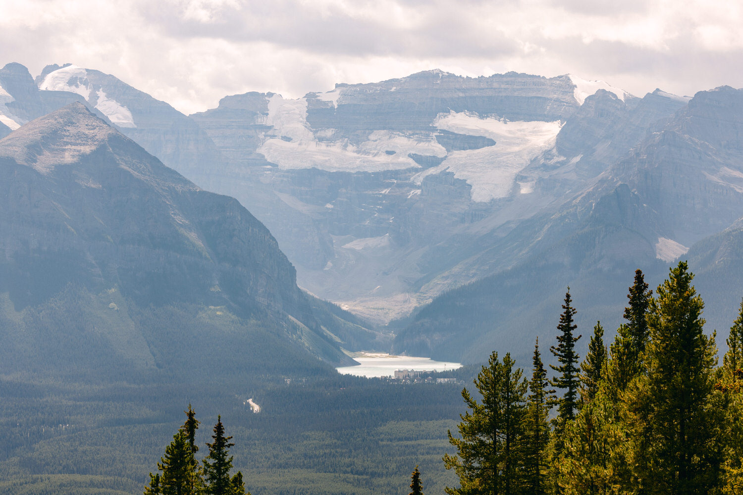 A scenic view of rugged mountains with glaciers and a serene lake nestled among dense forests under a cloudy sky.