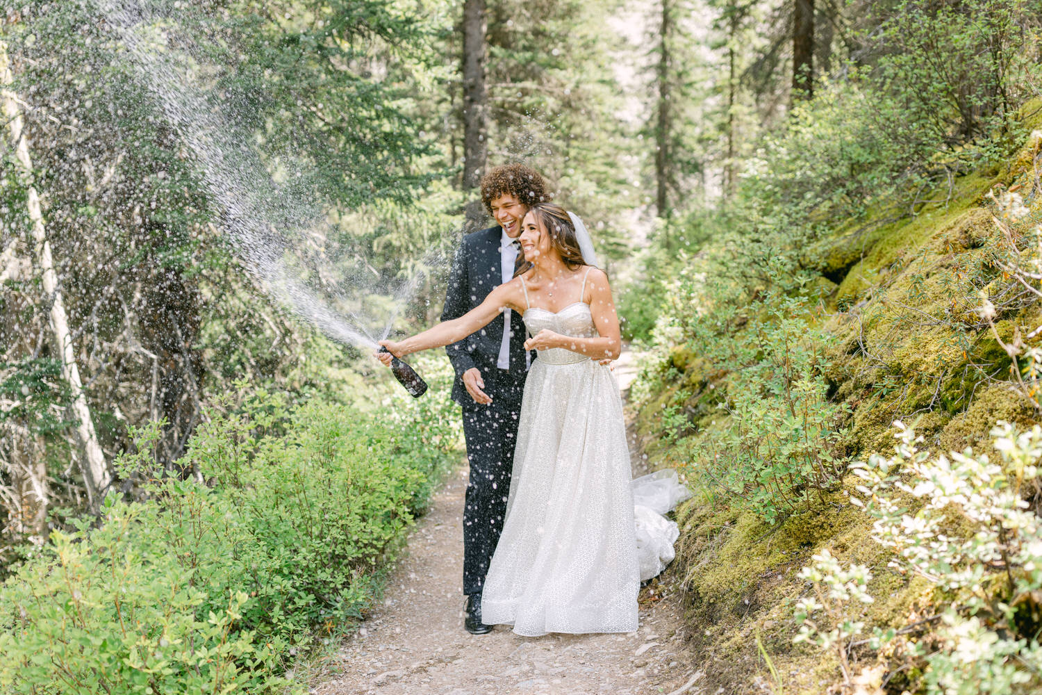 A couple joyfully sprays champagne in a lush forest during their wedding celebration.