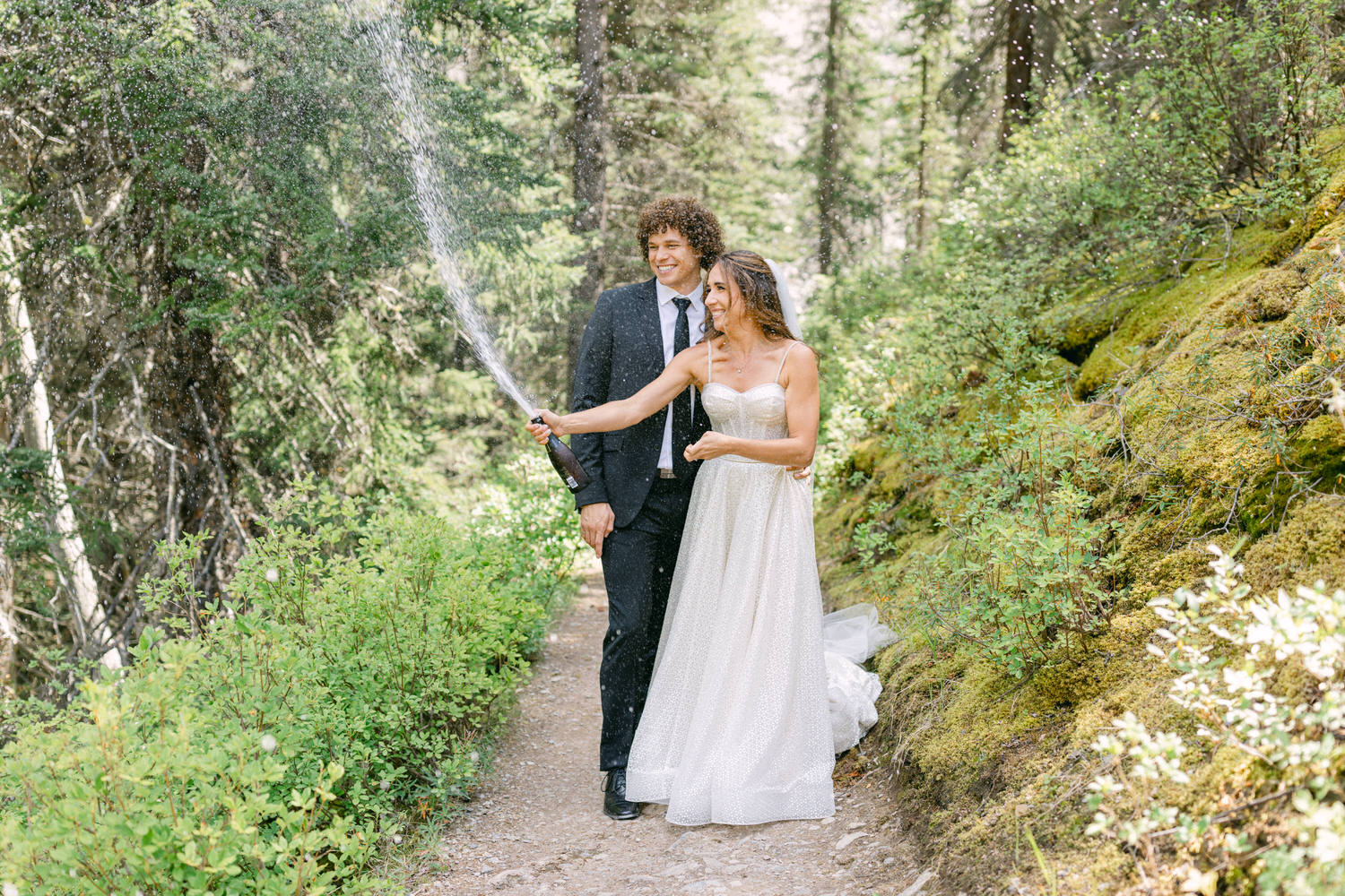 A joyful couple in formal wedding attire pops a champagne bottle in a lush, green forest, capturing a moment of celebration and happiness.