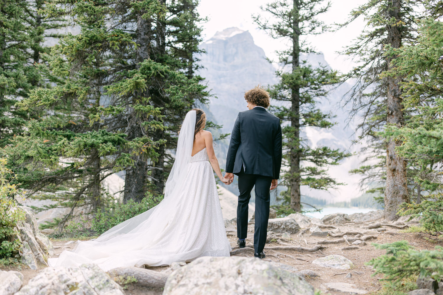 A bride and groom holding hands, walking through a forested path with mountains in the background.