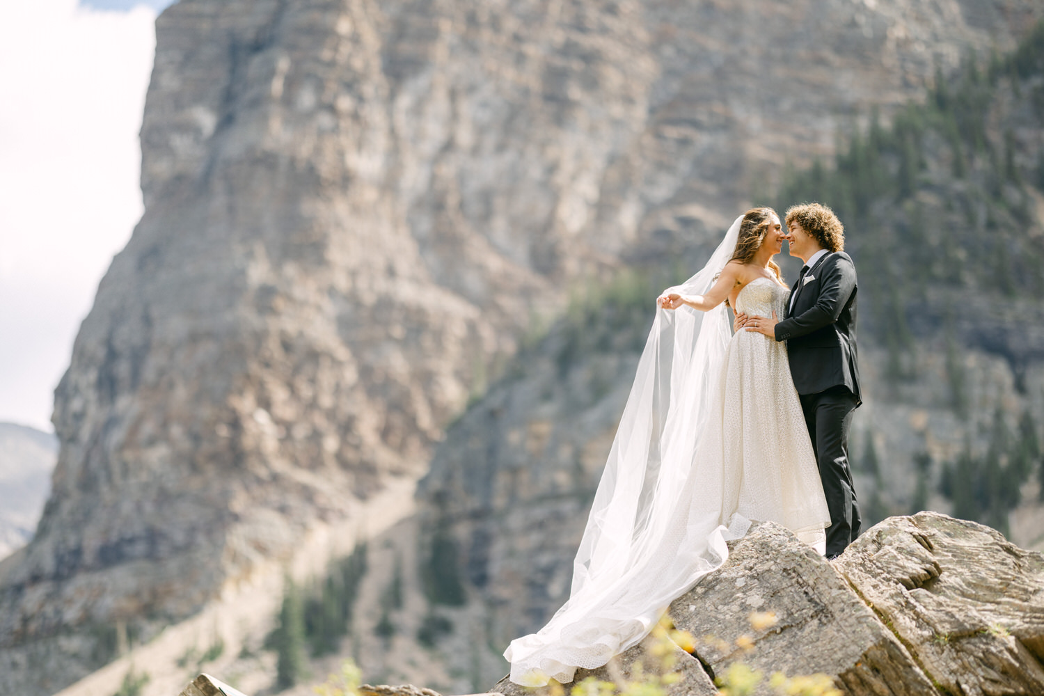 A couple joyfully embracing on a rocky outcrop with a scenic mountain backdrop. The bride wears a beautiful gown and veil, while the groom is in a classic suit.