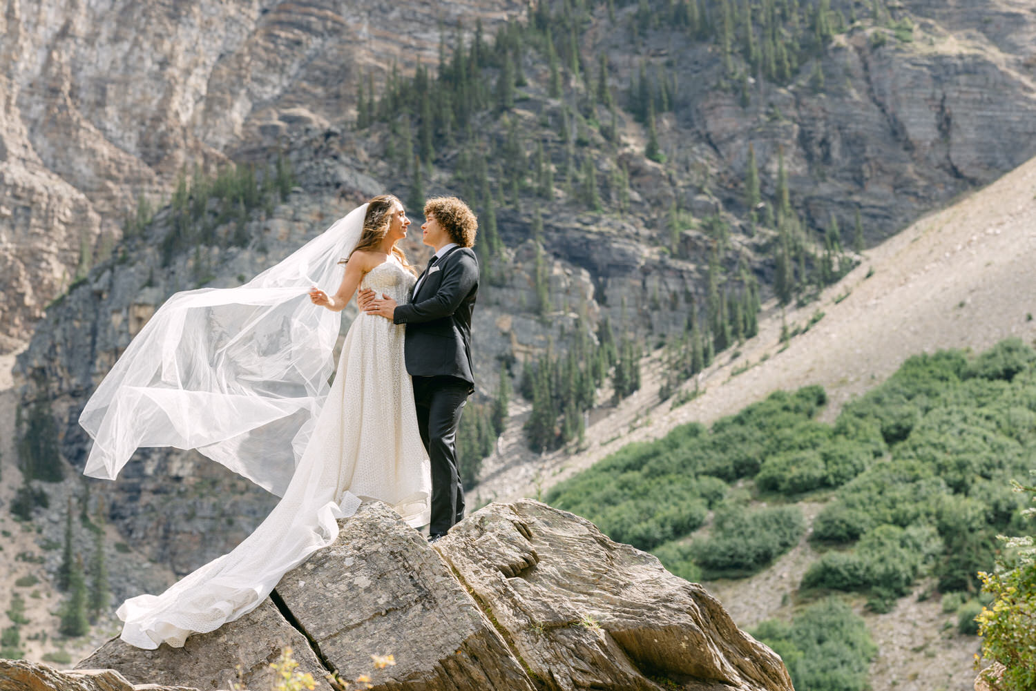 A couple sharing a romantic moment atop a rocky outcrop, surrounded by lush mountains, with the bride's veil gracefully flowing in the wind.