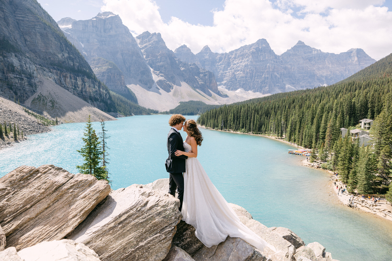 A bride and groom share a romantic moment on a rocky outcrop overlooking a turquoise lake and majestic mountains in the background.