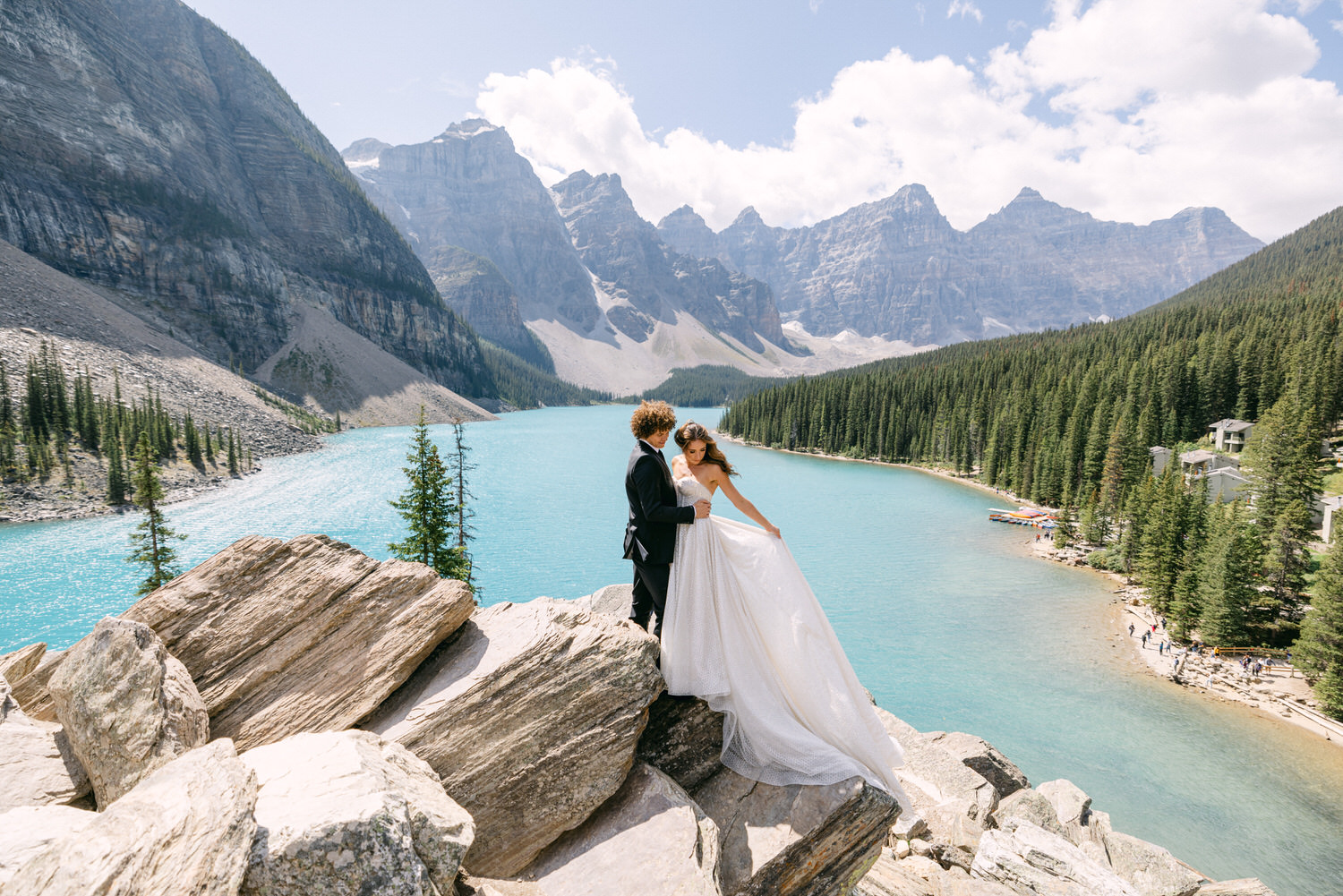 A couple in wedding attire stands on large rocks near a turquoise lake surrounded by mountains and pine trees, capturing a serene and picturesque moment.