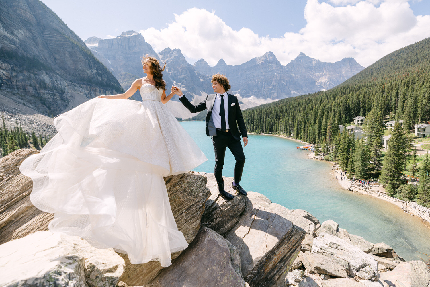 A couple in formal wedding attire stands on rocky terrain by a blue lake, surrounded by mountains and pine trees, celebrating their love amidst breathtaking scenery.