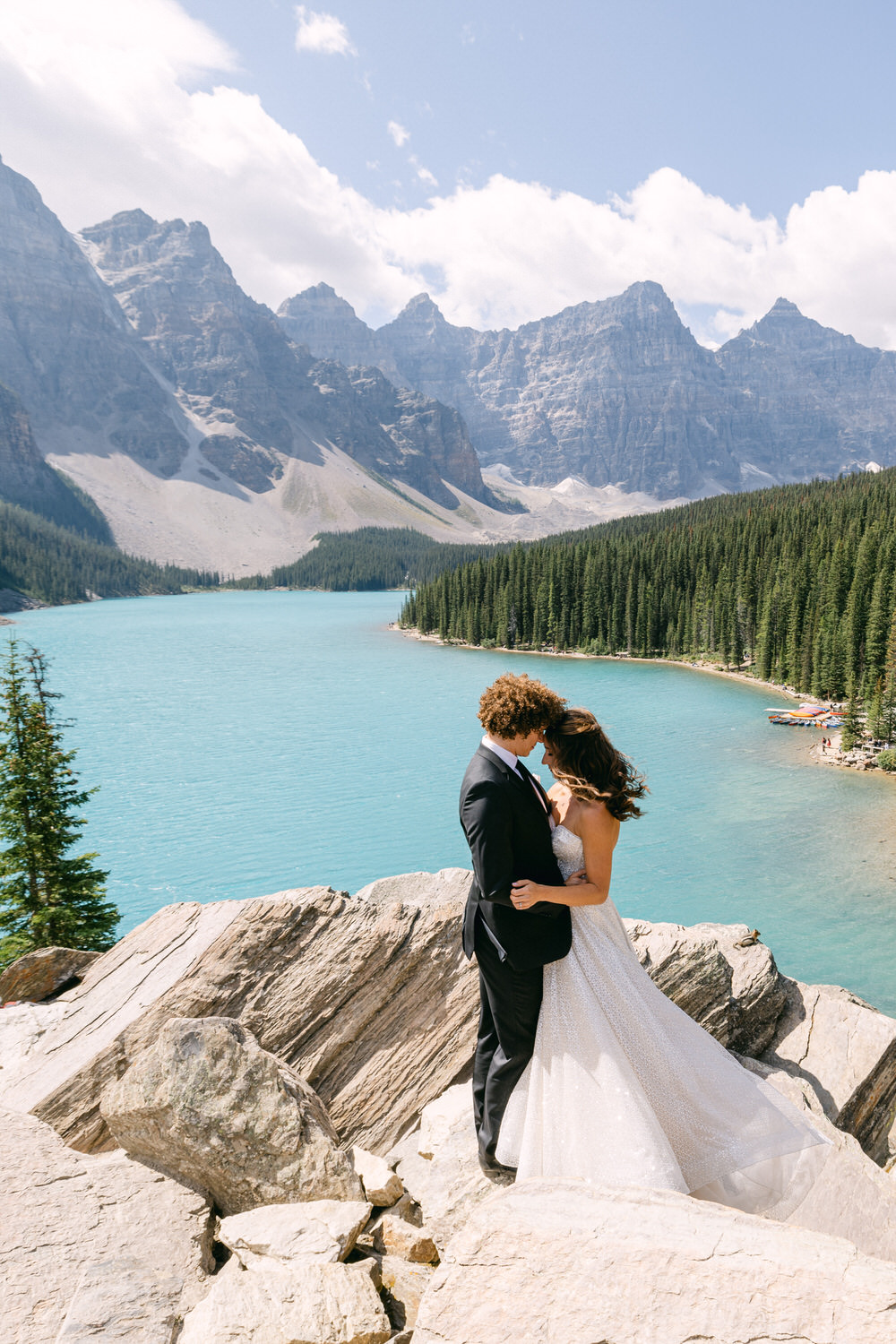 A couple embracing on rocky terrain by a turquoise lake, surrounded by towering mountains and lush forests.