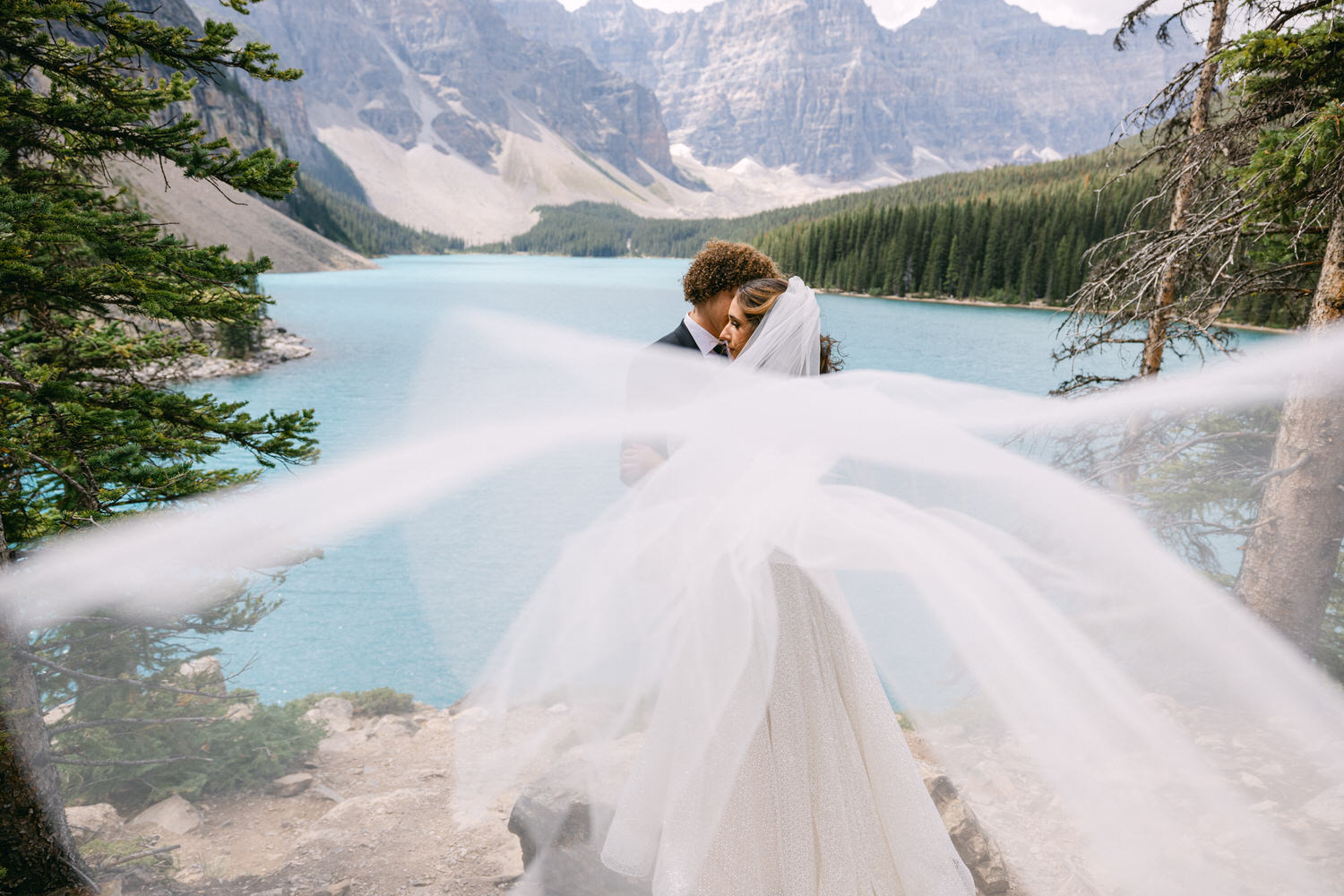 A couple embraces intimately against a stunning lake backdrop, with flowing veil and picturesque mountains.