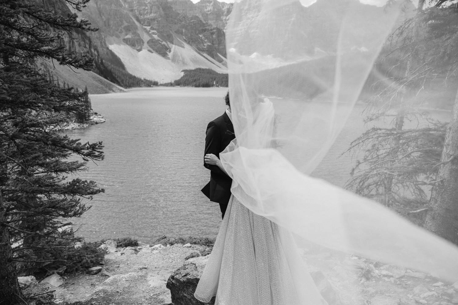 A couple shares a tender moment, partially obscured by a flowing veil, with a serene lake and mountains in the background.