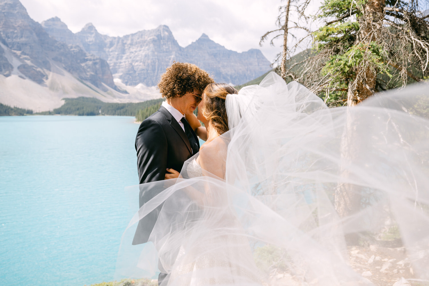 A couple in formal wedding attire sharing a romantic moment by a turquoise lake, surrounded by majestic mountains, with the bride's flowing veil capturing the breeze.