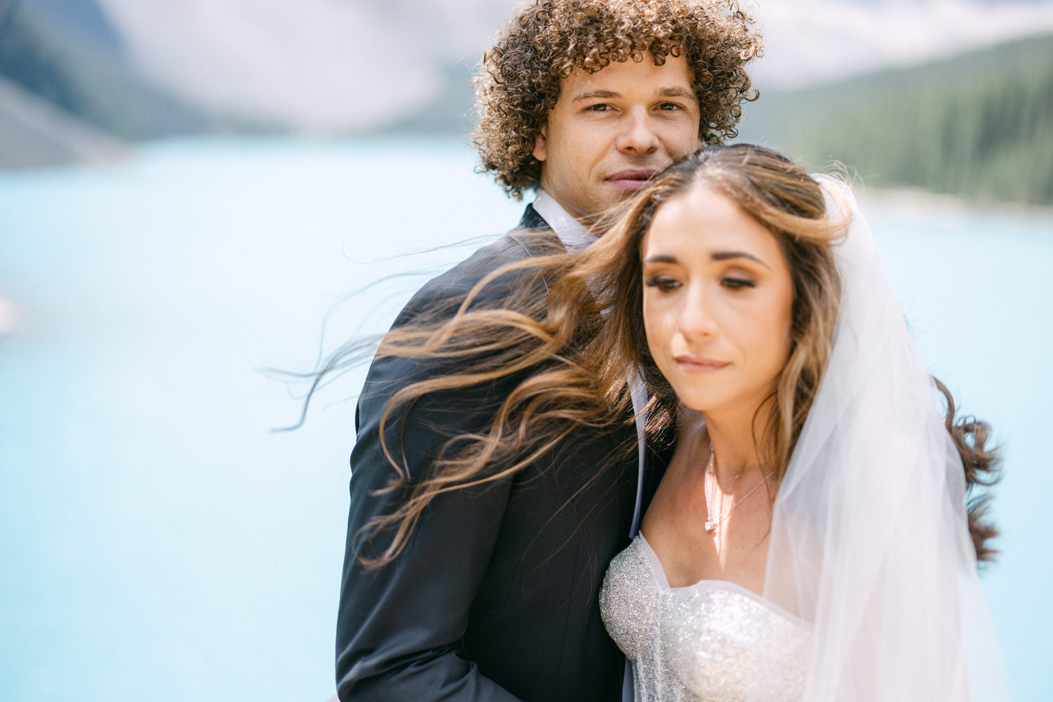 Couple embracing by a turquoise lake with mountains in the background, the bride wearing a sparkling gown and veil.