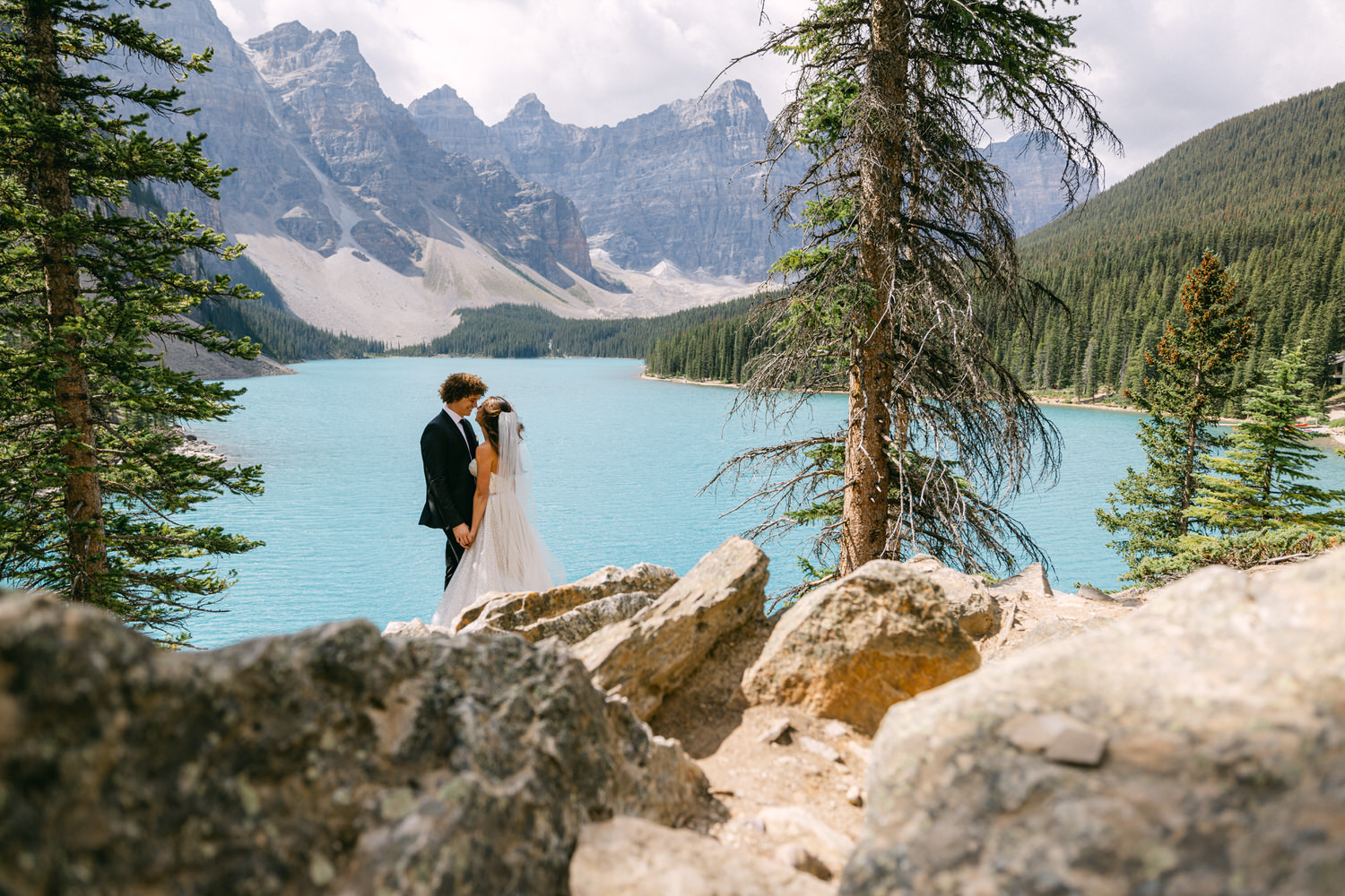 A bride and groom embrace in a picturesque outdoor setting by a turquoise lake, surrounded by mountains and trees.