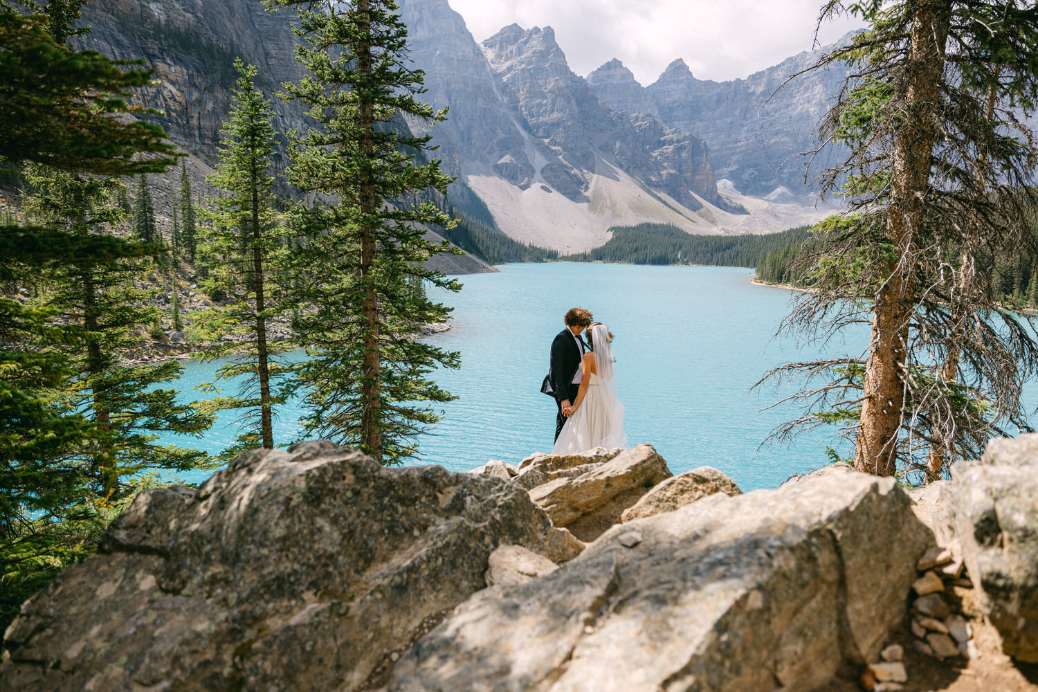 A couple embraces on a rocky outcrop with a stunning turquoise lake and mountains in the background, surrounded by evergreen trees.