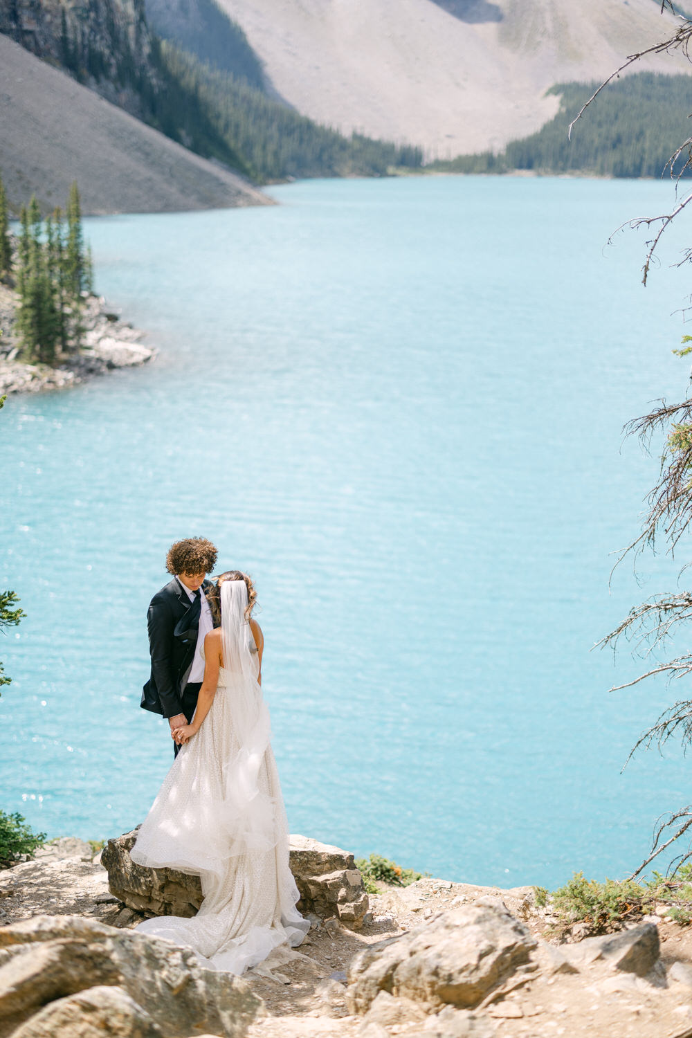 A couple holds hands in an intimate embrace by a stunning turquoise lake, surrounded by mountains and trees.