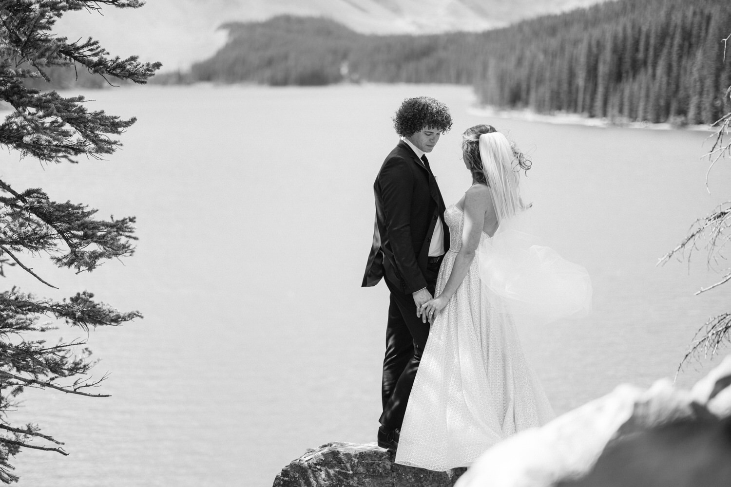 A couple holding hands on a rocky outcrop by a serene lake, surrounded by evergreen trees and mountains, captured in black and white.