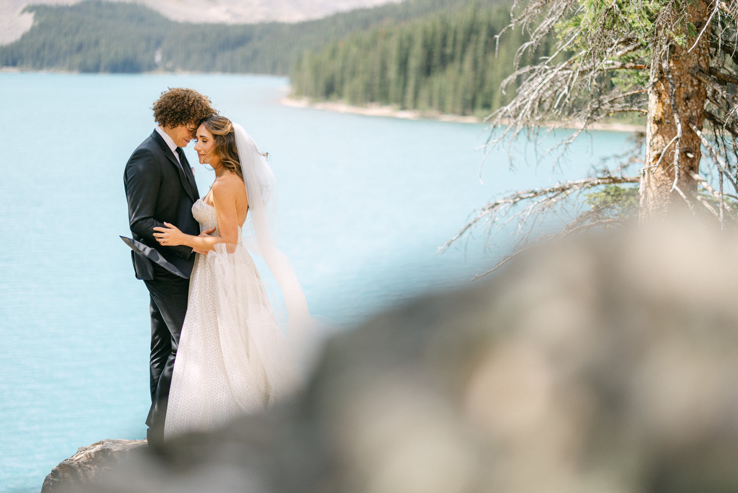 A couple sharing a tender moment by a turquoise lake, surrounded by nature, dressed in wedding attire.