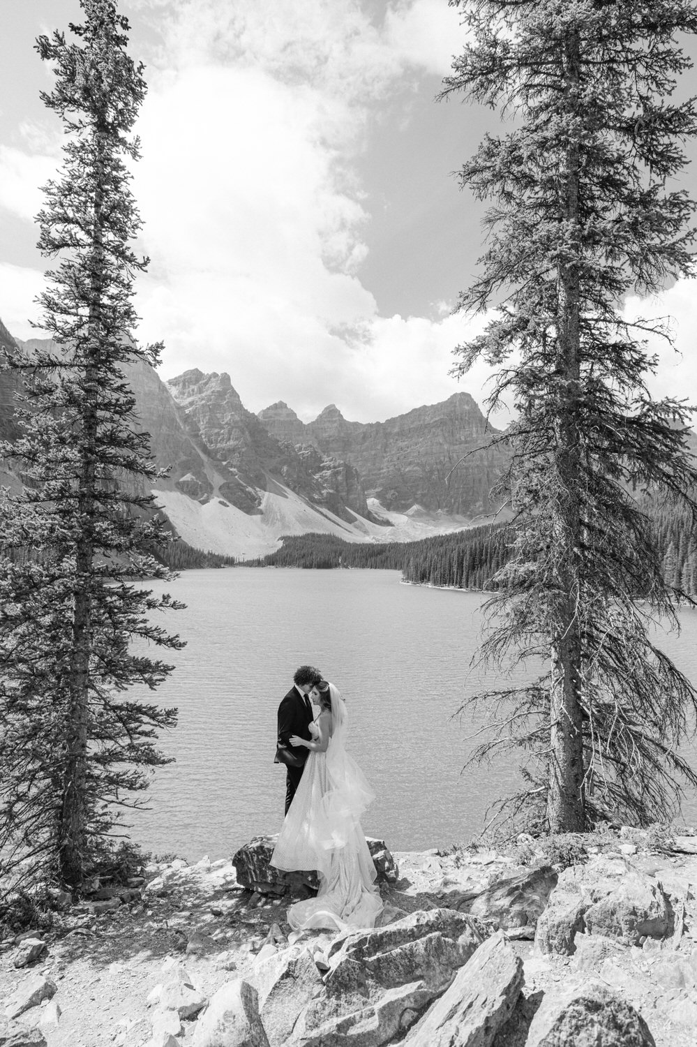 A couple in wedding attire shares a kiss by a serene lake surrounded by mountains and trees, captured in black and white.