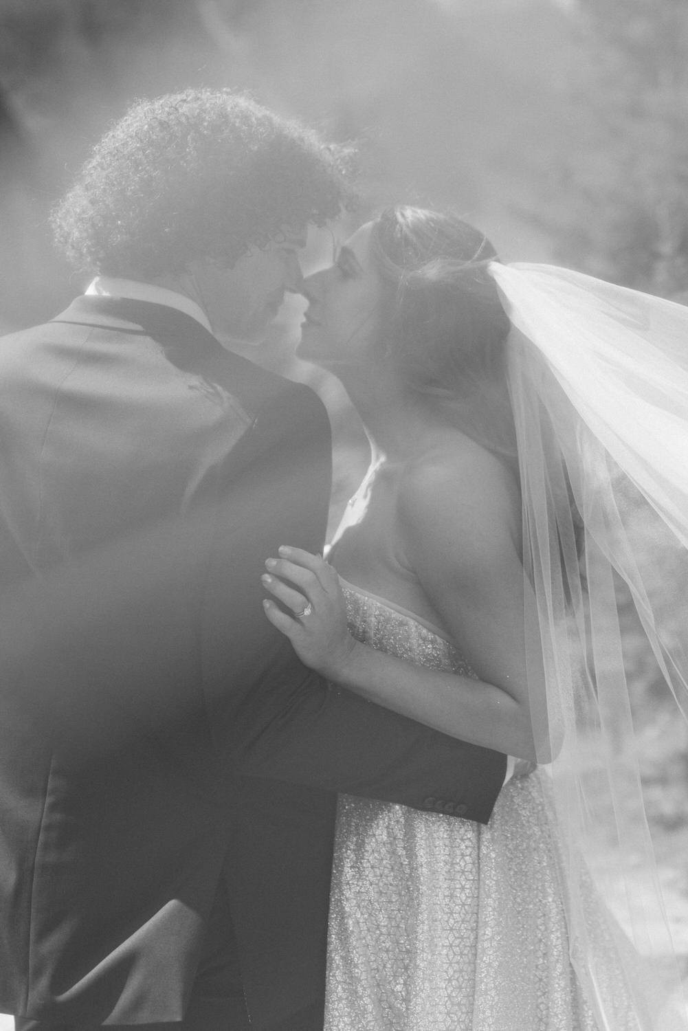 A couple shares a tender kiss, captured in soft focus with a romantic atmosphere, showcasing the bride in a sparkling dress and groom in a classic suit.