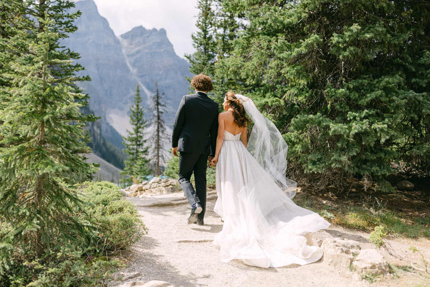 A bride and groom walk hand-in-hand along a scenic pathway surrounded by trees and mountains, with the bride's flowing veil and dress trailing behind her.