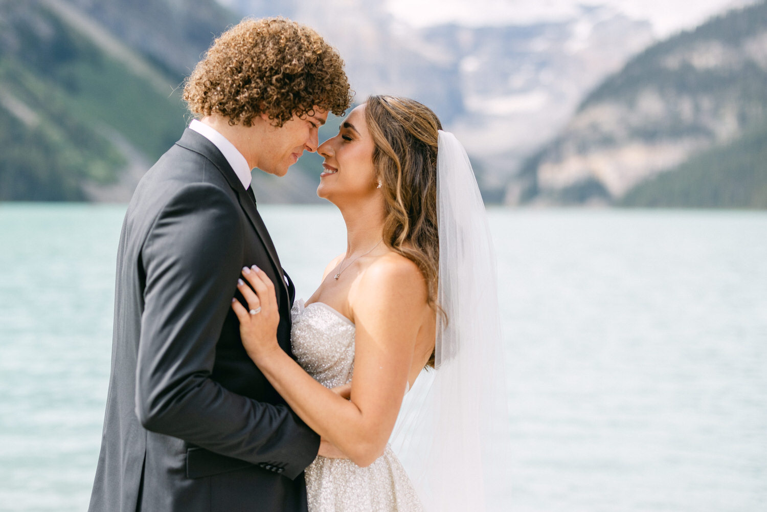 A couple shares a loving gaze by a scenic lake, surrounded by mountains, during their wedding ceremony.