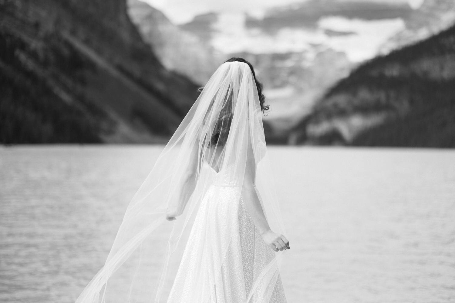 A bride in a flowing white dress and veil stands by a serene lake, with mountains in the background, captured in black and white.