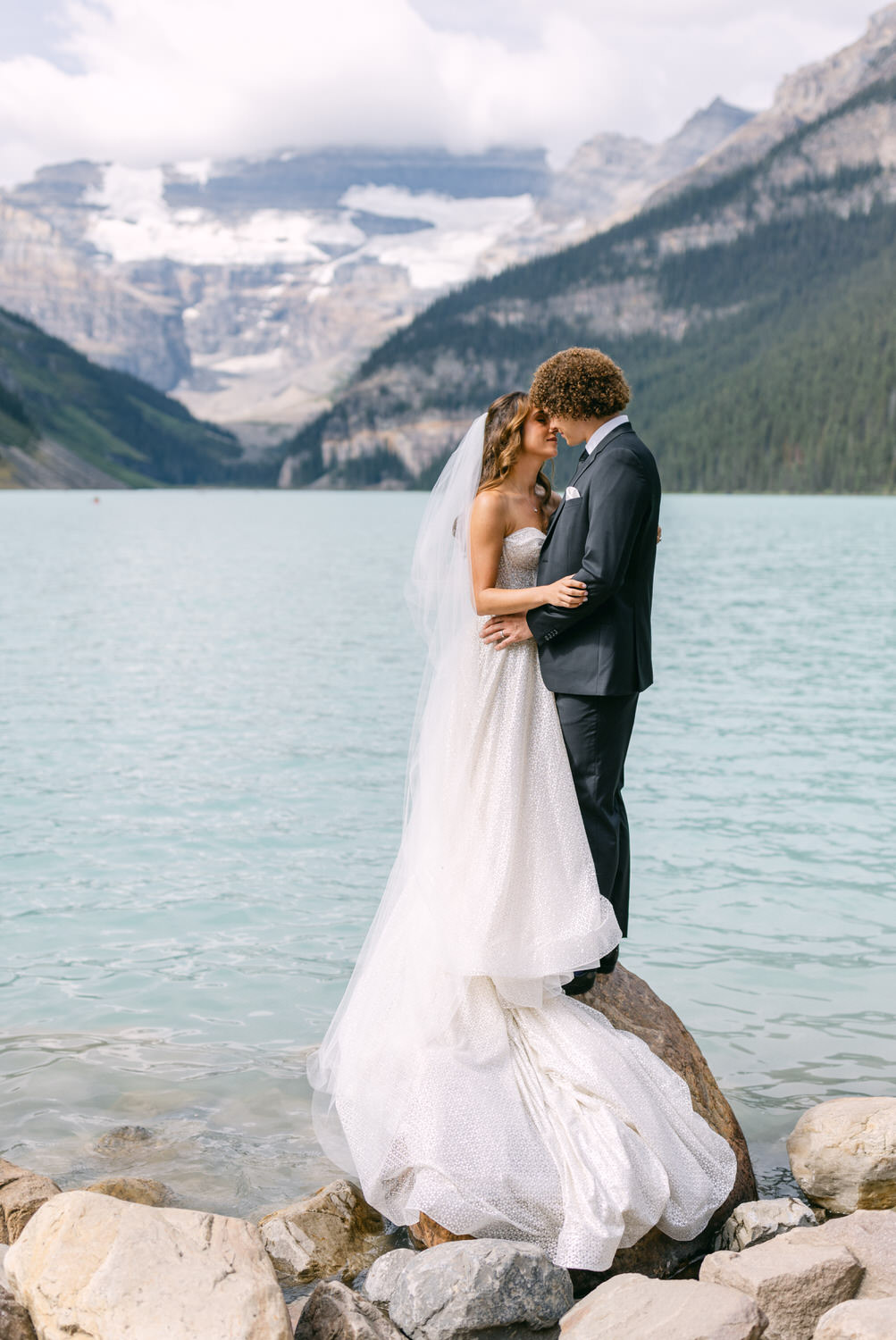 A bride and groom share a tender kiss while standing on a rock by a serene lake, surrounded by mountains and greenery.