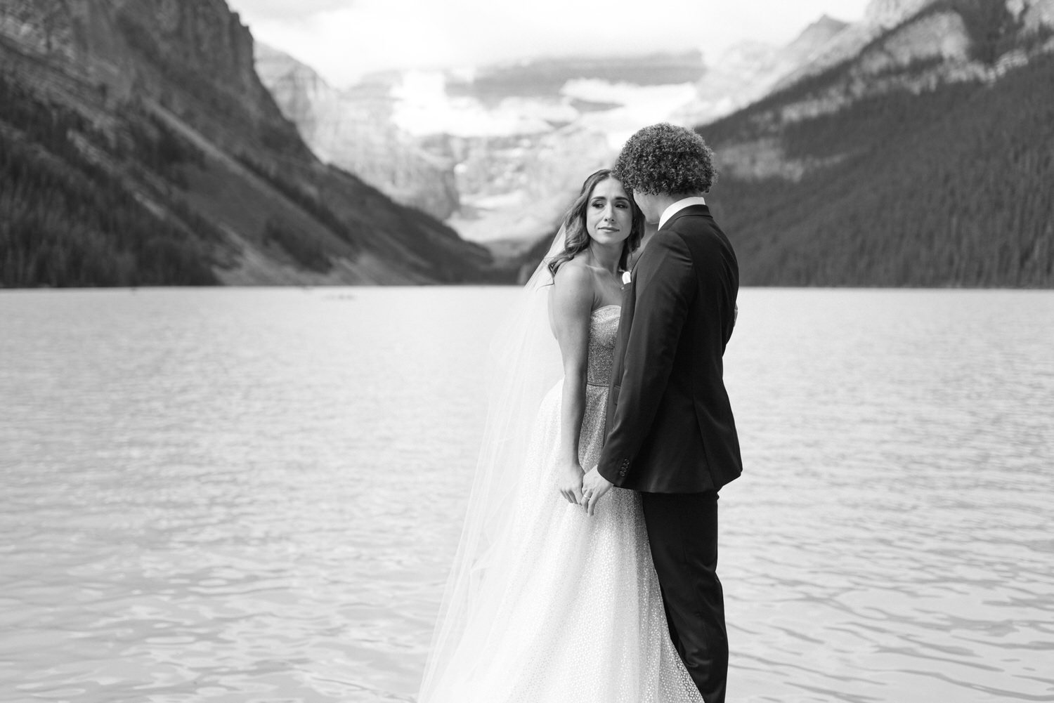 A bride and groom share a tender moment by the serene waters of Lake Louise, surrounded by mountains in black and white.