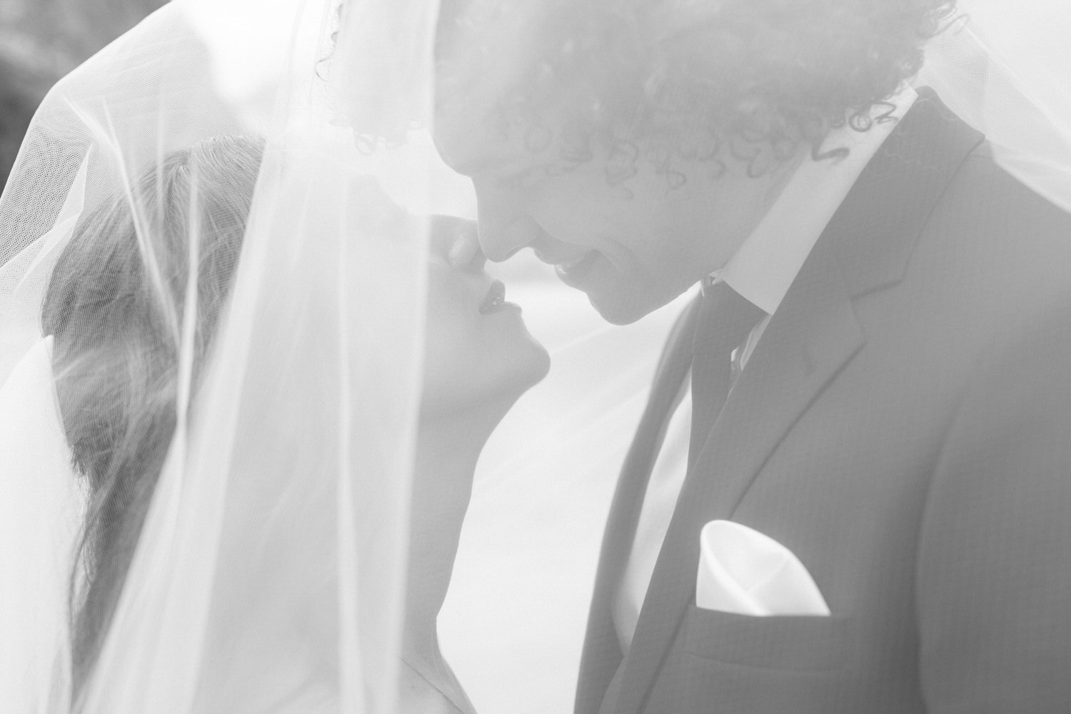 A close-up of a couple gazing into each other's eyes under a veil, capturing a romantic moment in black and white.