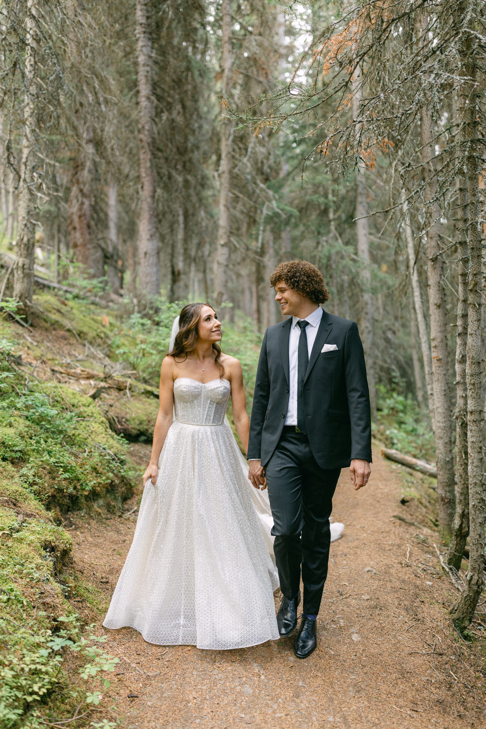 A bride in a sparkling gown and a groom in a suit walk hand-in-hand along a forest path, surrounded by tall trees and lush greenery, sharing a joyful glance.