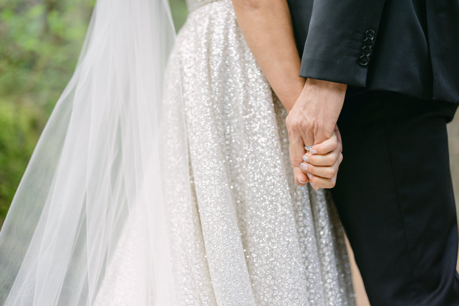 A close-up of a couple holding hands, showcasing their wedding rings and the bride's shimmering dress, with a soft veil in the background.