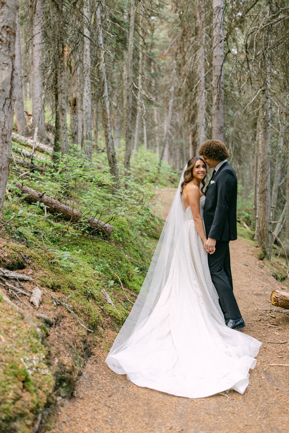 A bride and groom share a joyful moment in a serene forest, holding hands amidst tall trees and lush greenery, with the bride's flowing veil and gown creating a romantic scene.