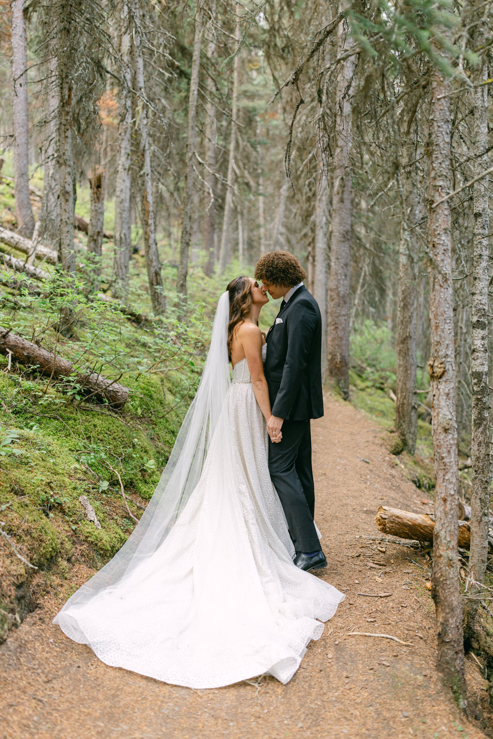 A bride and groom share a kiss on a wooded trail, surrounded by tall trees and lush greenery, capturing a serene and intimate moment.