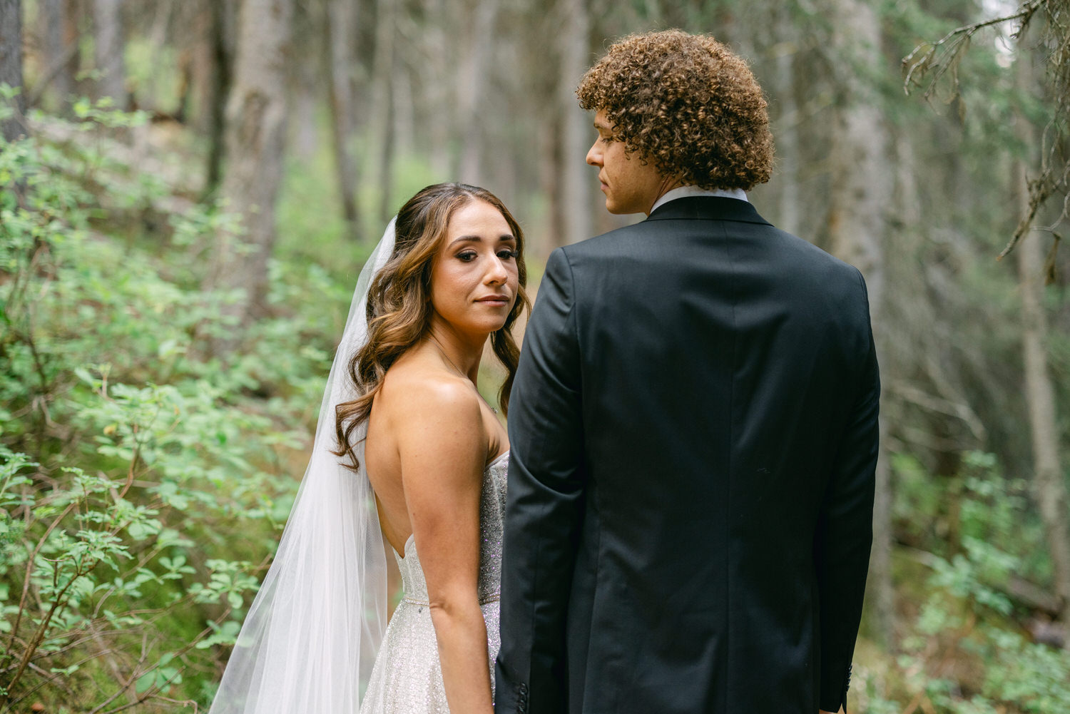 A bride and groom pose together in a serene forest setting, the bride looking back at the camera while the groom faces away.