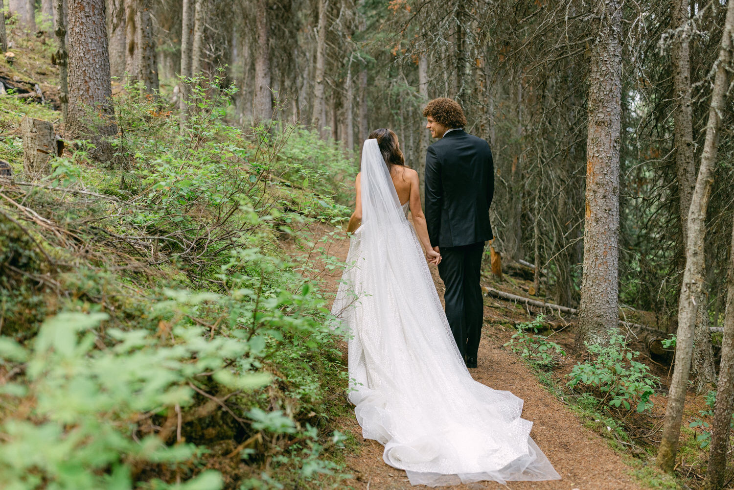 A couple holding hands walks down a wooded path, adorned in wedding attire, surrounded by lush greenery and tall trees.