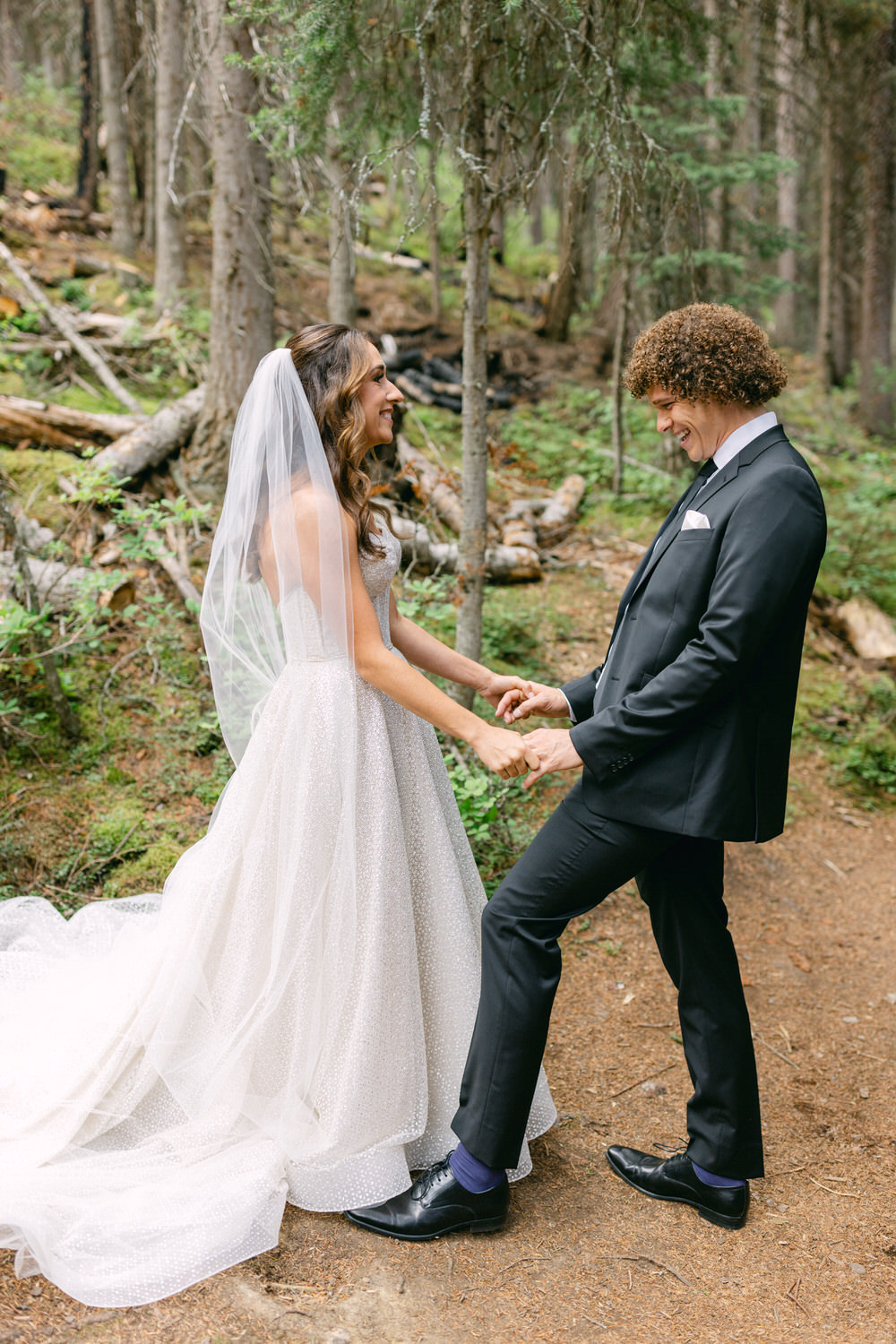 A couple shares a joyful moment in a forest clearing during their wedding ceremony, with the bride in a sparkling gown and veil, and the groom in a classic black suit.