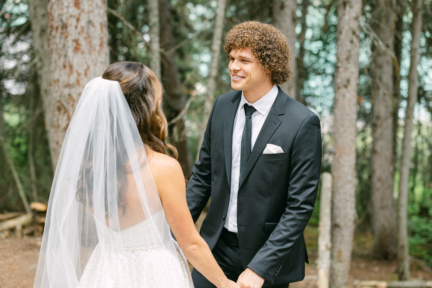 A groom in a black suit smiles at his bride in a sparkling wedding dress and veil, surrounded by lush trees.