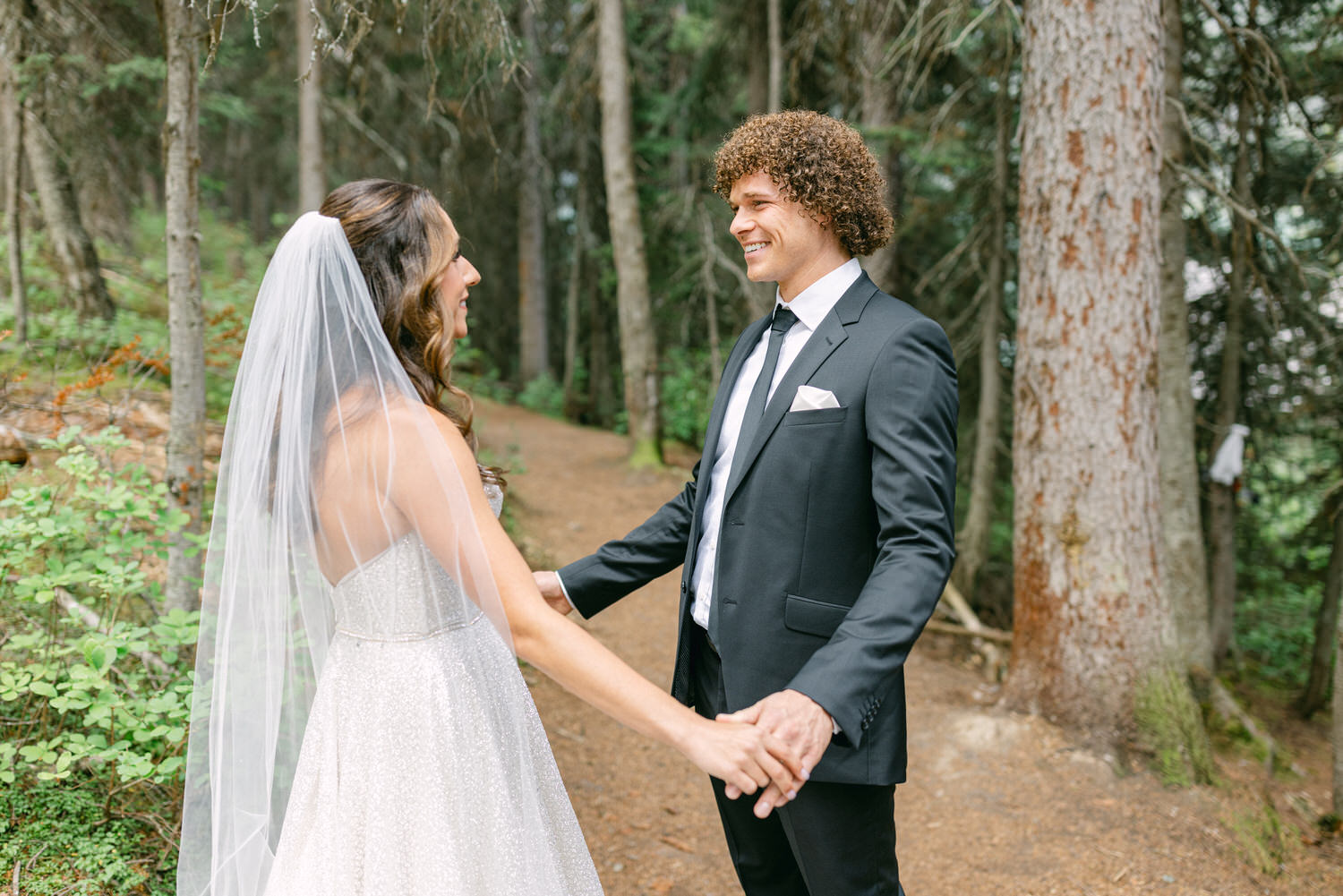 A bride and groom holding hands and smiling at each other in a forest setting surrounded by tall trees.