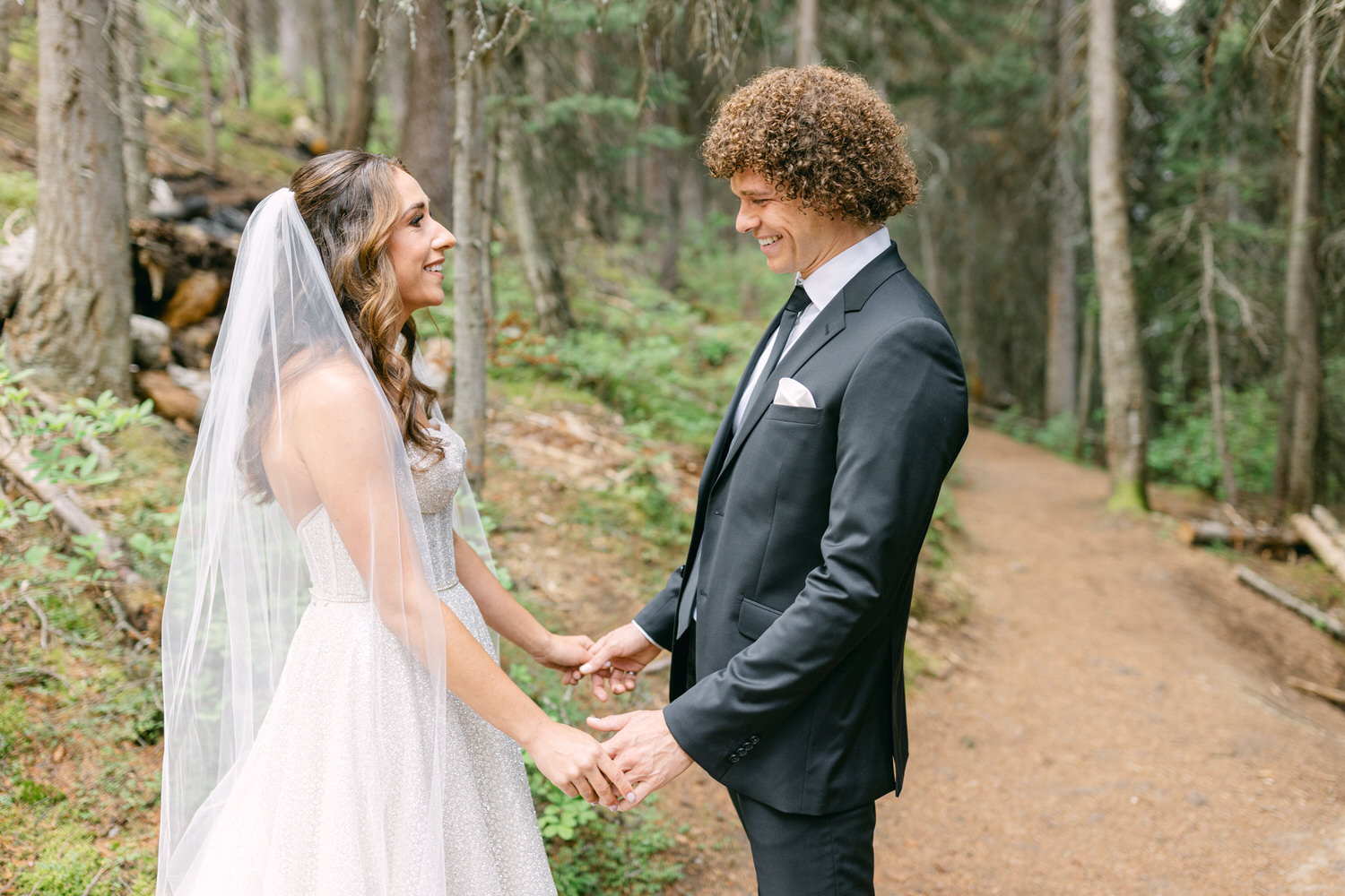 A bride and groom hold hands and share a moment of joy in a lush forest, surrounded by tall trees and greenery.