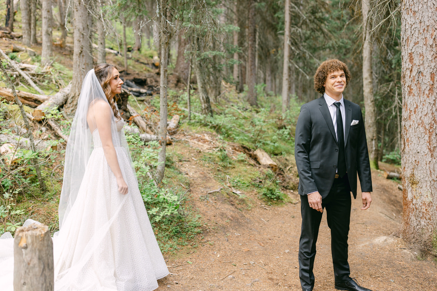 A bride in a flowing gown and veil stands nearby as the groom, dressed in a black suit, faces away with a smile, surrounded by a serene forest backdrop.