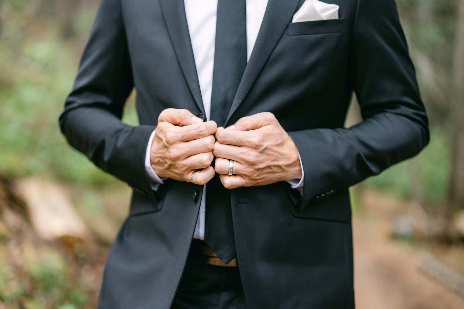 Close-up of a groom fastening his black suit jacket in a forest setting, showcasing wedding attire and subtle wedding rings.