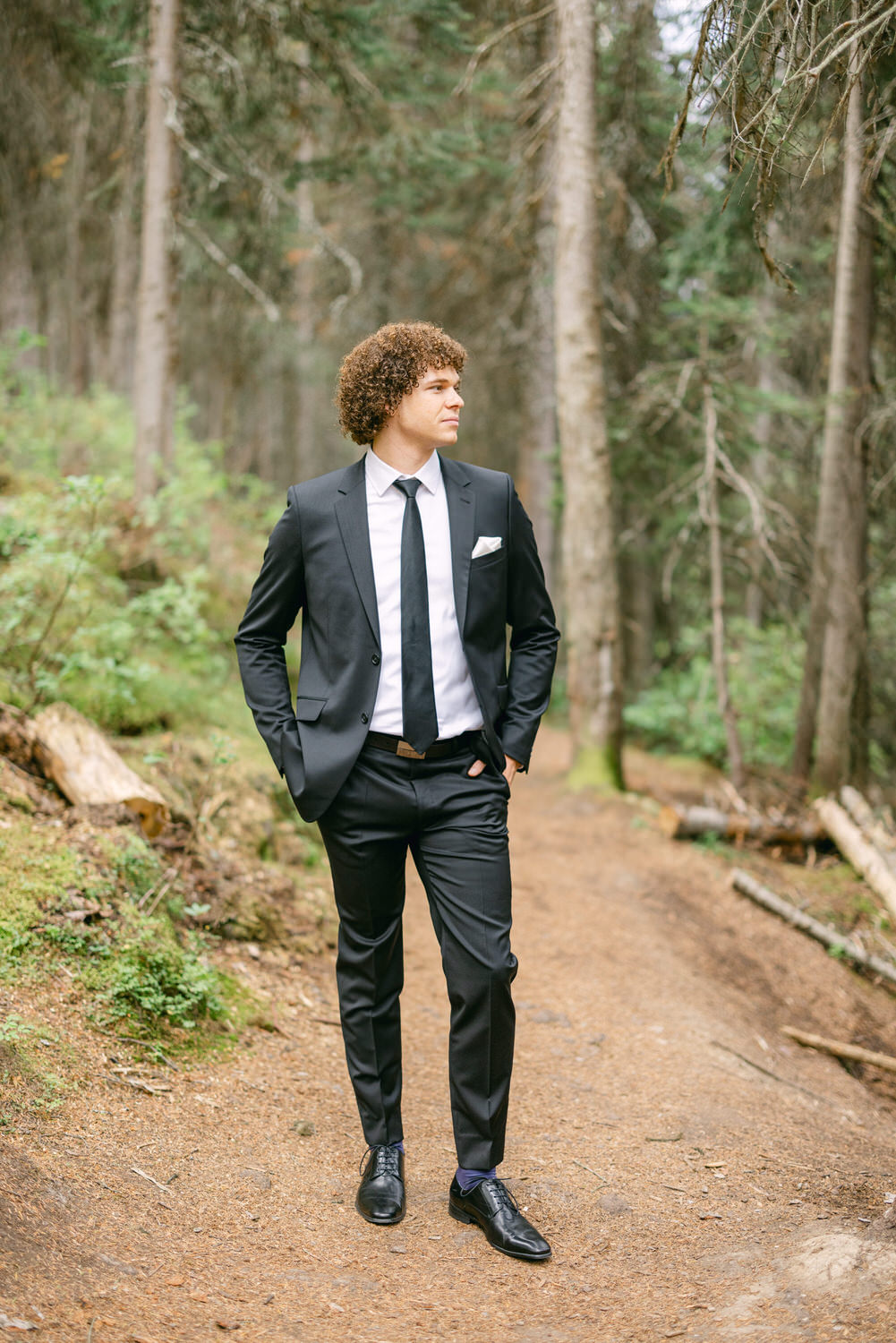 A well-dressed man with curly hair walks along a wooded path, wearing a black suit, white shirt, and black tie, surrounded by lush greenery and tall trees.