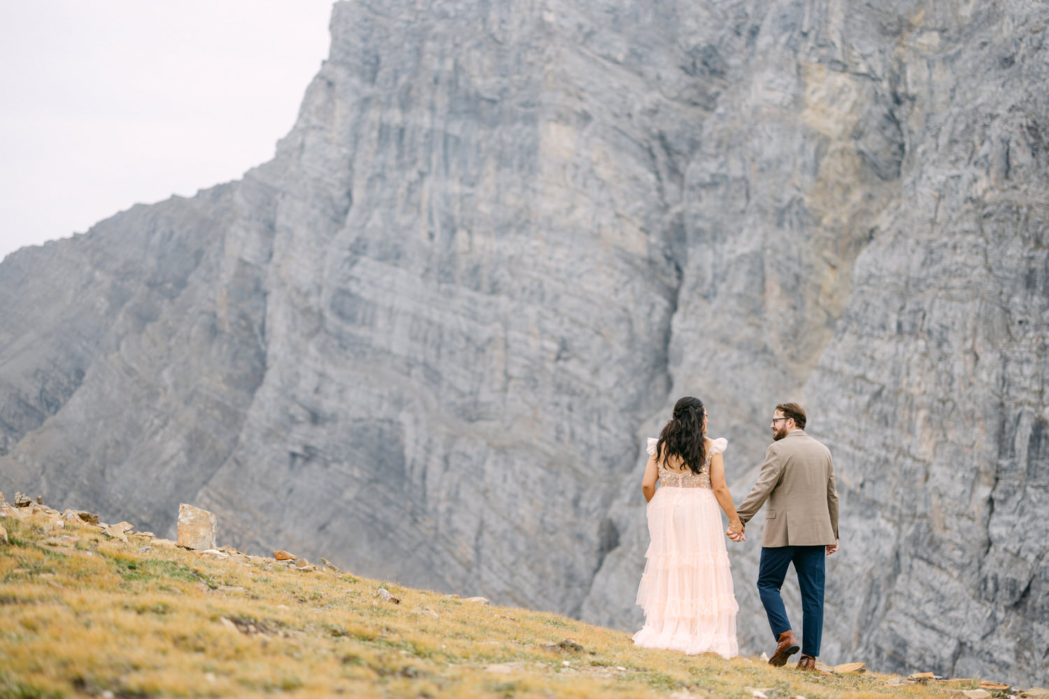 A couple holds hands while walking on a grassy area with a rugged mountain backdrop, dressed in elegant attire.