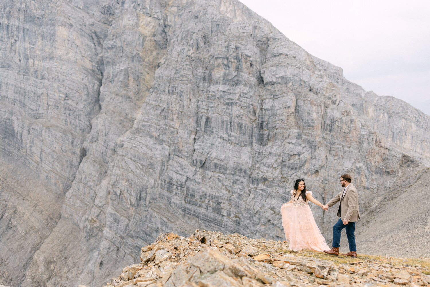 A couple holds hands on a rocky mountaintop, dressed in elegant attire against a backdrop of towering grey mountains.