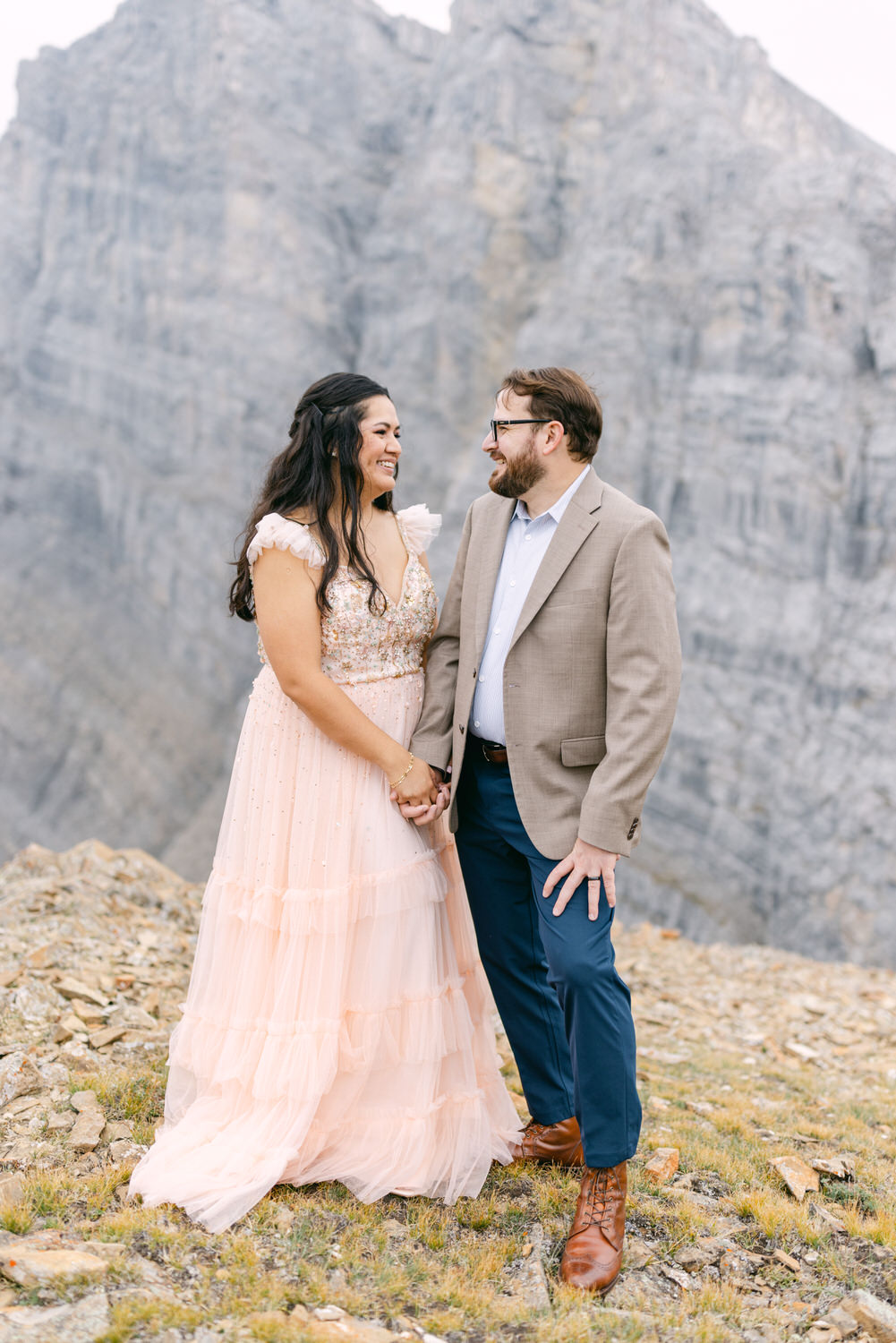 A couple shares a joyful interaction while holding hands, surrounded by a stunning mountainous backdrop. The woman is dressed in a beautiful pink gown with sequins, and the man is in a light blazer and glasses, both smiling at each other.