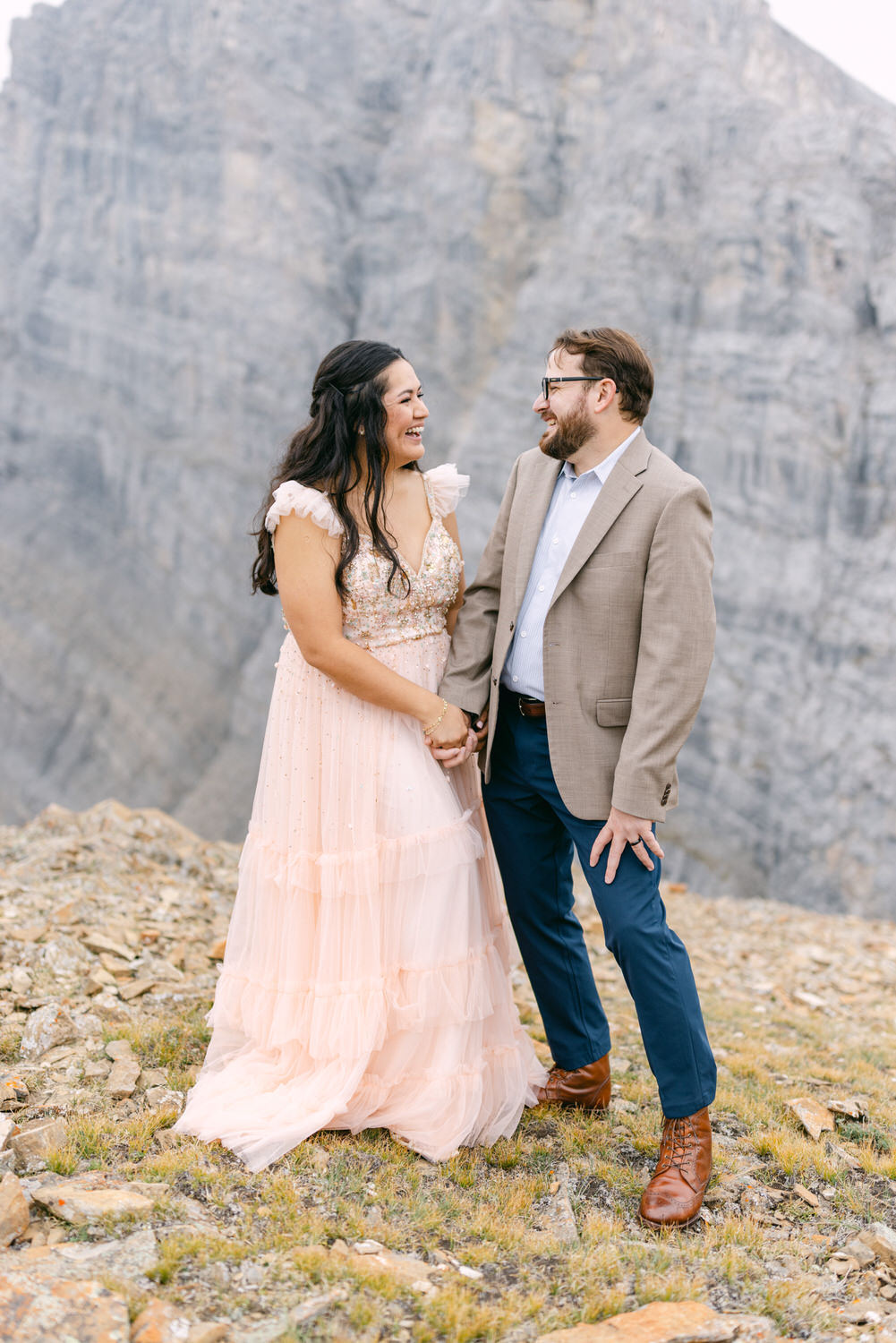 A smiling couple holds hands outdoors, with a stunning rocky landscape in the background; the woman is dressed in a flowing pink gown, and the man wears a blazer and glasses.